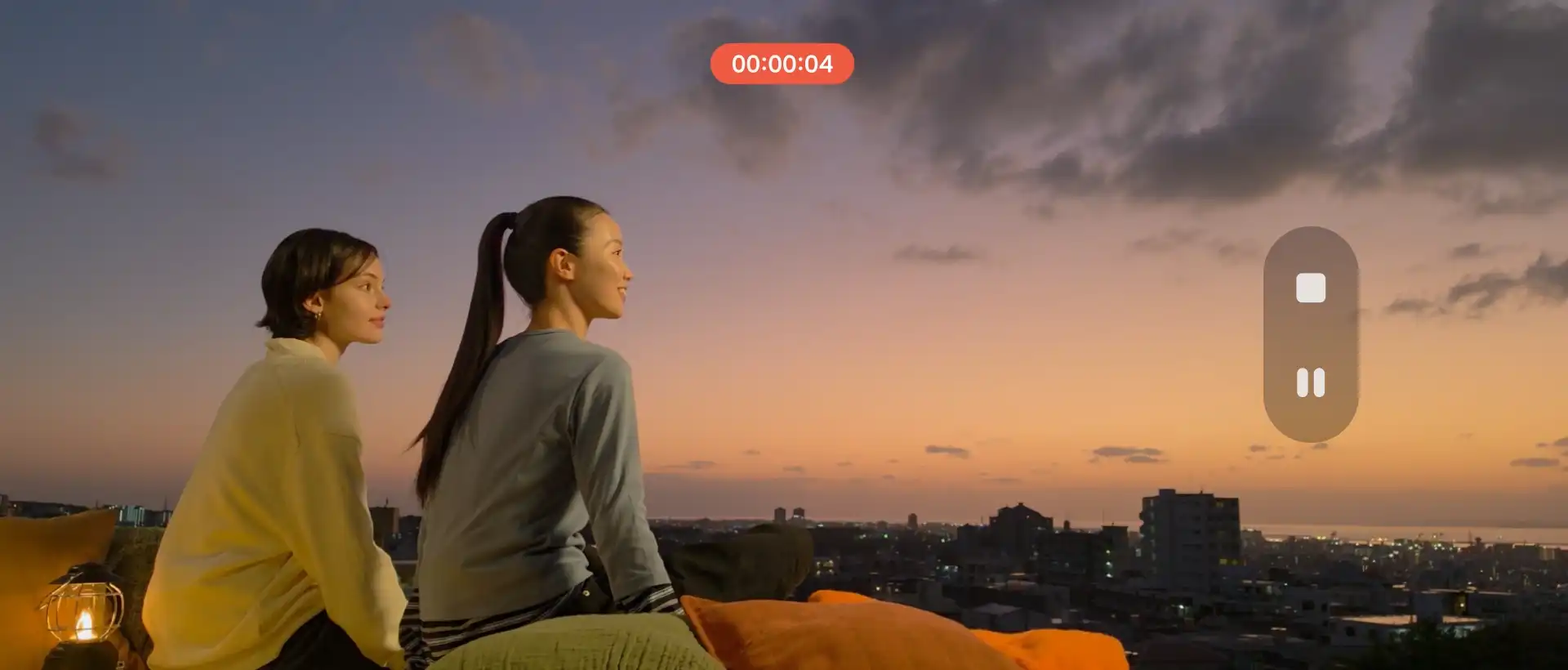 Két nő naplementét néz a város felett, a kijelzőn videófelvétel kezelőfelülete látható.