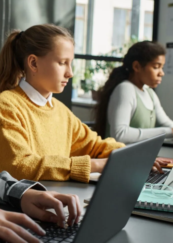 Diákok ülnek egy tanteremben és laptopon dolgoznak; az előtérben egy sárga pulóveres lány látható munka közben. Digitális oktatás, tanulás, iskola, technológia az oktatásban, diákok, laptop, tantermi munka, jövő oktatása.