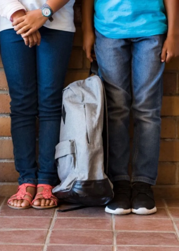 Két farmernadrágos gyermek áll egy téglafal előtt, közöttük egy szürke hátizsák hever a földön. Iskolakezdés, barátság, gyerekek, iskolatáska, hátizsák, tanévkezdés, hétköznapi pillanat.