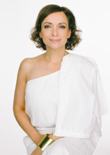 Portré egy mosolygós, barna hajú nőről, aki fehér ruhát visel, valószínűleg görög stílusú, félvállas drapériával. A nő rövid, hullámos barna hajjal rendelkezik, és enyhén mosolyogva néz a kamerába.