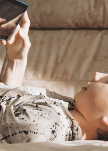 Egy kisfiú fekszik egy világos színű kanapén vagy ágyon, és egy tableten néz valamit. A kisfiú arca részben árnyékban van, a fény felülről, jobbról érkezik. A tablet kék kerettel rendelkezik, és a kisfiú enyhén megemelt karral tartja, közben háton fekszik