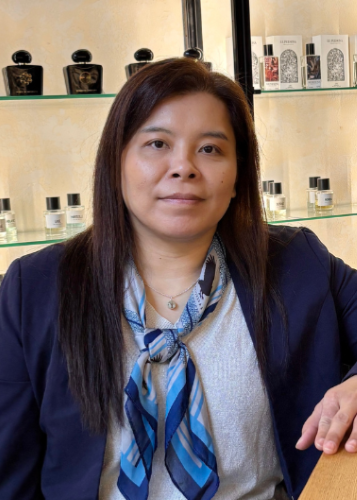 Sötét hajú nő sötétkék zakóban és kék mintás sállal egy parfüméria polcai előtt, amelyeken elegáns parfümös üvegek sorakoznak. Parfüméria, illatszerbolt, szakértő, üzlet belső, elegáns nő, kozmetikumok, luxus termékek.