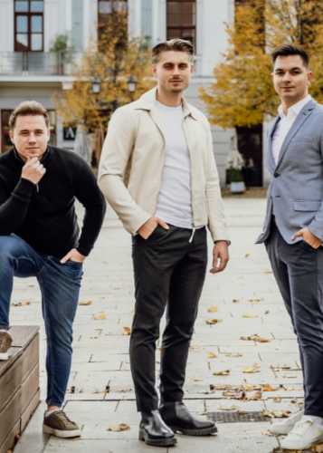 Három fiatal férfi áll egy őszi városi téren, modern öltözékben. Férfiak, város, divat, barátok, őszi hangulat, elegancia.