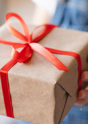 Közelkép egy barna nátronpapírba csomagolt ajándékról, amelyet fényes piros szalag és masni díszít. Az ajándékot egy személy tartja, akinek csak a kezei és ruhájának egy része látszik a háttérben, jelezve, hogy az ajándékot átadják vagy kapják. 