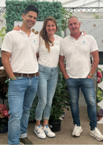 A Gardencentrum háromtagú csapata – két férfi és egy nő – fehér pólóban és kék farmerben mosolyog a kamerába egy kiállításon.